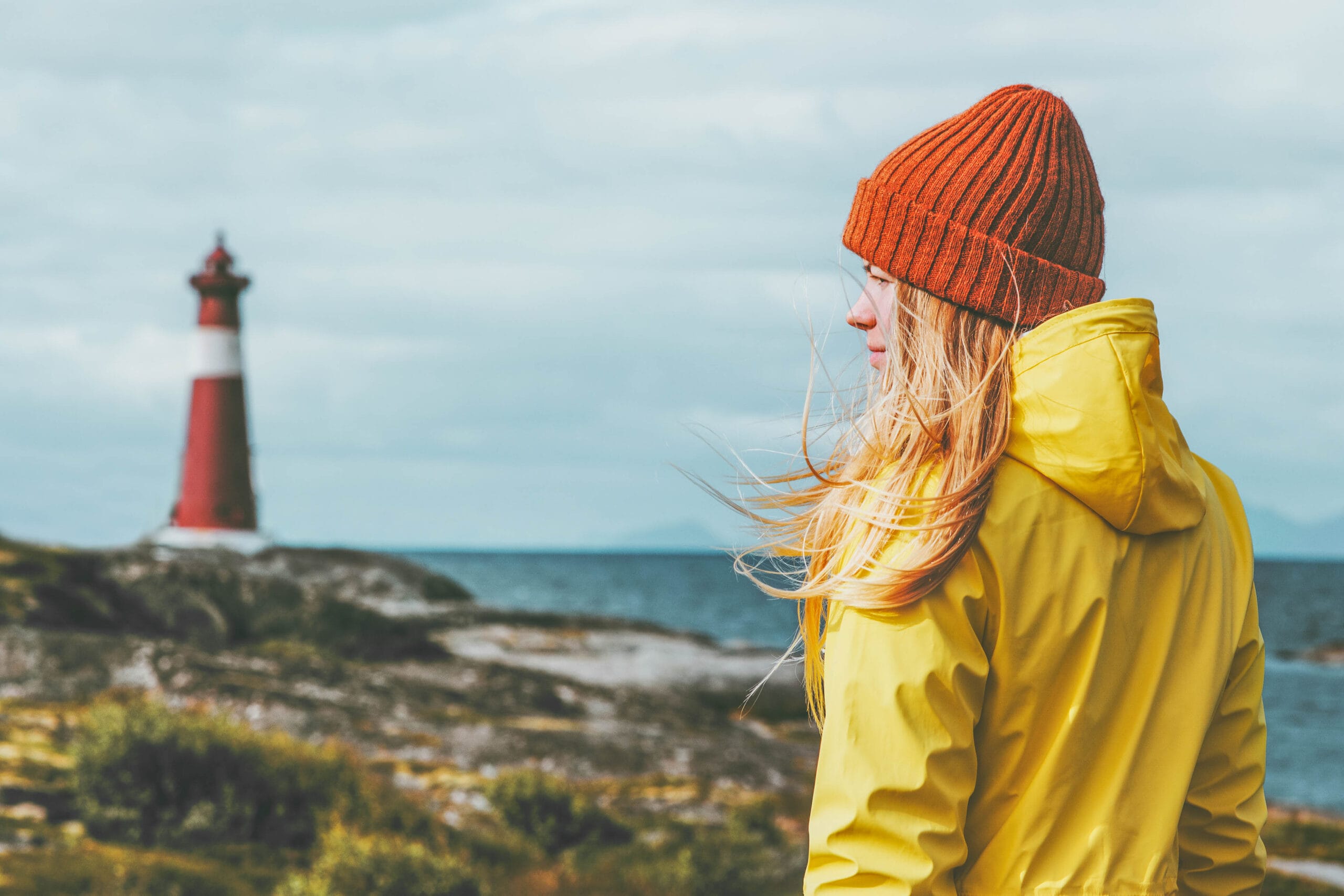Kvinne med gul regnjakke og lue ved havet og et fyr