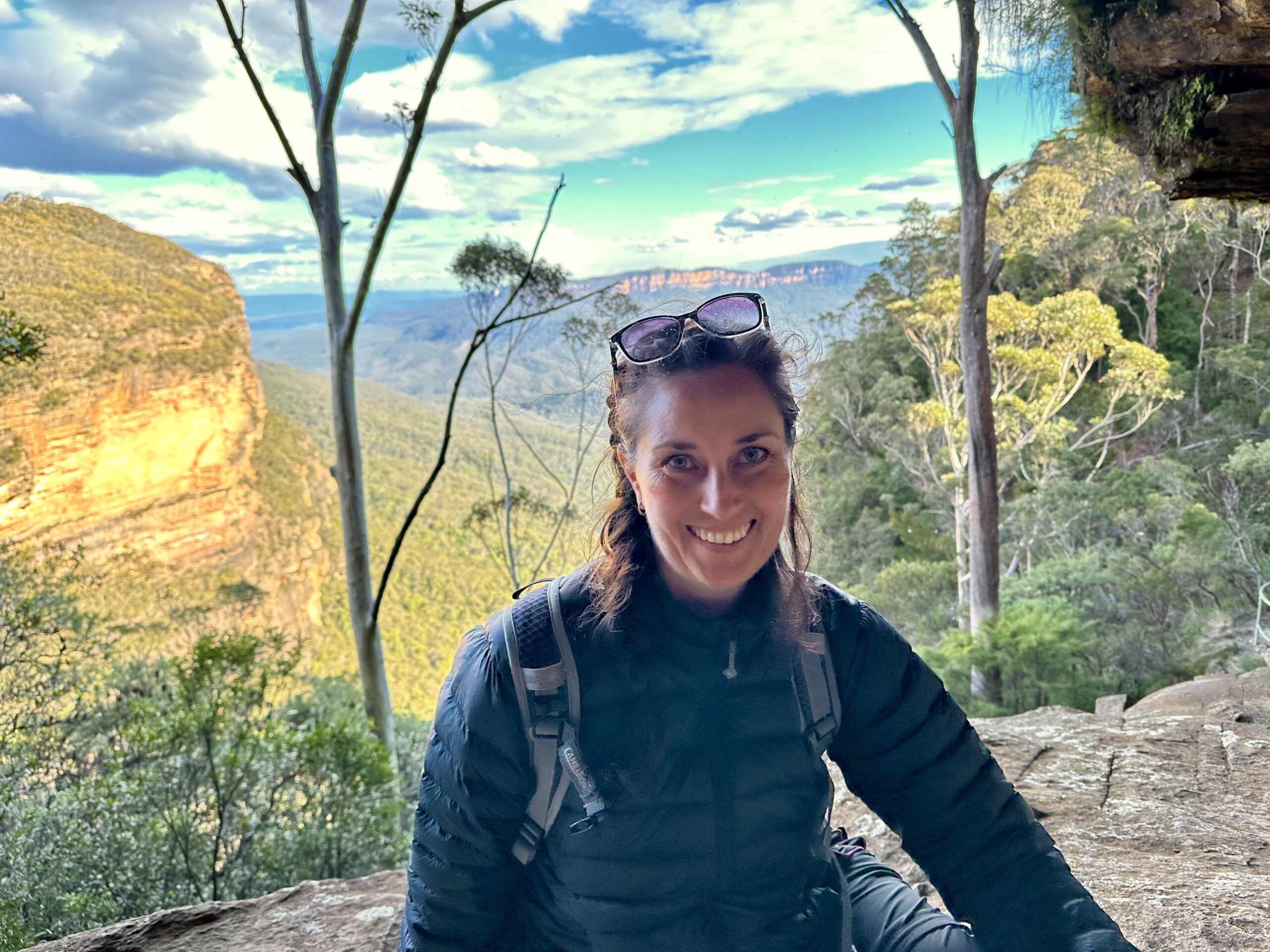 Smiling woman in a dark jacket stands on a rocky overlook with a canyon, trees and a blue sky in the background, sunglasses on her head.