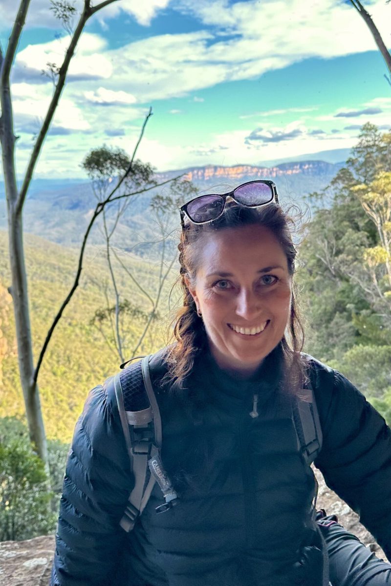 Smiling woman in a dark jacket stands on a rocky overlook with a canyon, trees and a blue sky in the background, sunglasses on her head.